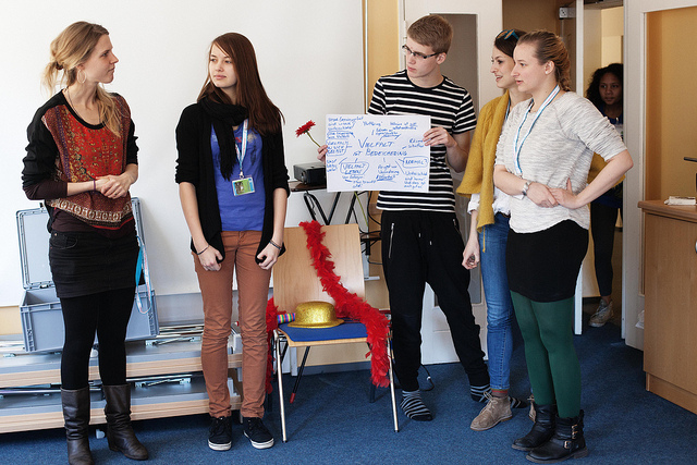 KI generiert: Das Bild zeigt eine Gruppe von fünf Personen in einem Innenraum, die sich zu unterhalten scheinen. Im Vordergrund steht ein Mann mit einem selbstgemachten Schild, während die anderen Personen in einer lockeren Runde um ihn herumstehen.