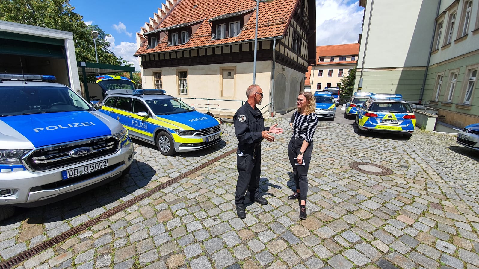 KI generiert: Das Bild zeigt eine Szene auf einem gepflasterten Platz mit mehreren Polizeiwagen im Hintergrund. Im Vordergrund unterhalten sich ein Polizist und eine Frau.