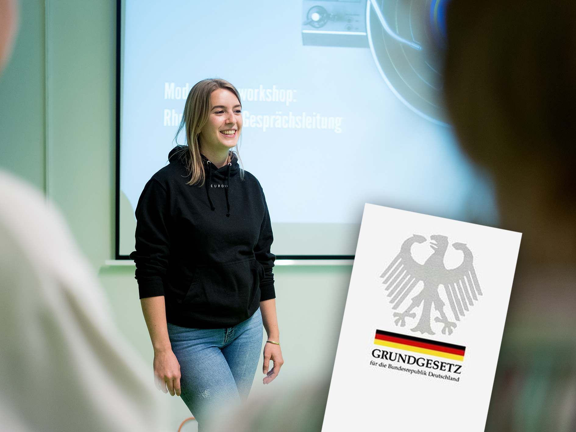 KI generiert: Das Bild zeigt eine lächelnde Person in einem Hoodie, die vor einem Projektor steht, auf dem der Titel eines Workshops zu sehen ist. Im Vordergrund ist das Logo des Grundgesetzes der Bundesrepublik Deutschland zu erkennen.