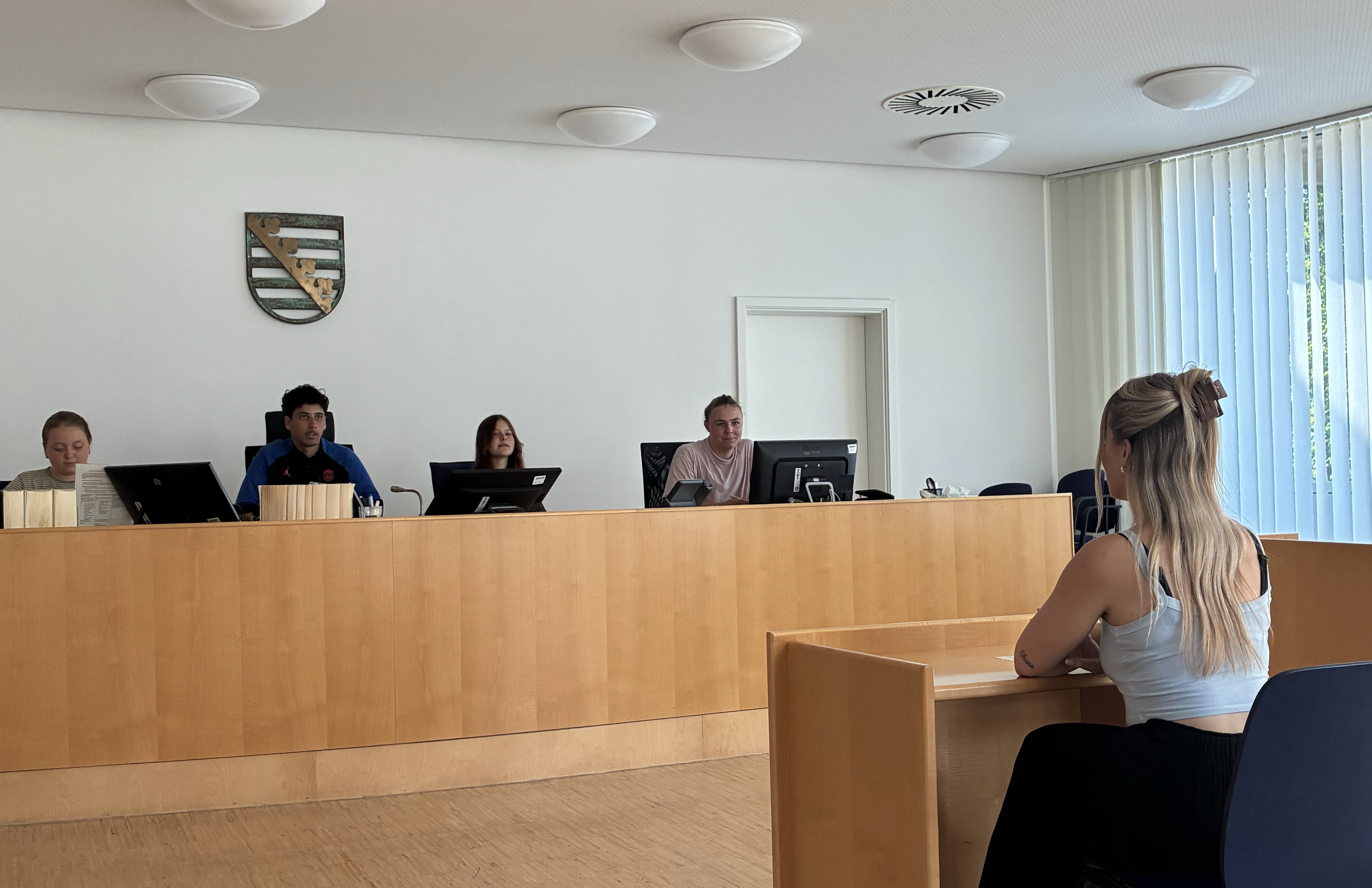 KI generiert: Das Bild zeigt einen Gerichtssaal mit mehreren Personen, darunter eine Frau, die am Tisch sitzt, und vier weitere Personen, die an einem erhöhten Tisch mit Computern arbeiten. Der Hauptinhalt des Bildes ist eine Gerichtssitzung oder eine ähnliche offizielle Anhörung.