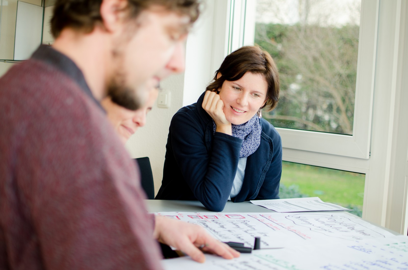 KI generiert: Das Bild zeigt zwei Personen, die an einem Tisch sitzen und sich auf ein Blatt Papier konzentrieren, das mit Notizen und Zeichnungen bedeckt ist. Der Hauptinhalt des Bildes ist eine Arbeits- oder Brainstorming-Sitzung.