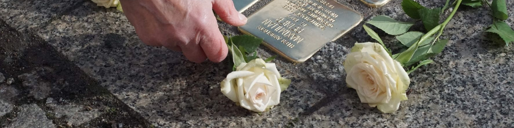KI generiert: Das Bild zeigt eine Hand, die eine goldene Gedenktafel auf einem Stein berührt, umgeben von zwei weißen Rosen. Der Hauptinhalt des Bildes ist eine Hommage oder Erinnerung an eine Person oder ein Ereignis, wie durch die Gedenktafel und die Blumen symbolisiert wird.