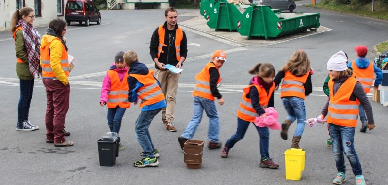 KI generiert: Das Bild zeigt eine Gruppe von Kindern und Erwachsenen, die orange Warnwesten tragen und sich auf einer Straße bewegen, während sie verschiedene Abfallbehälter mit sich führen. Der Hauptinhalt des Bildes scheint eine Aufräumaktion oder ein Umweltprojekt zu sein, an dem die Gruppe teilnimmt.