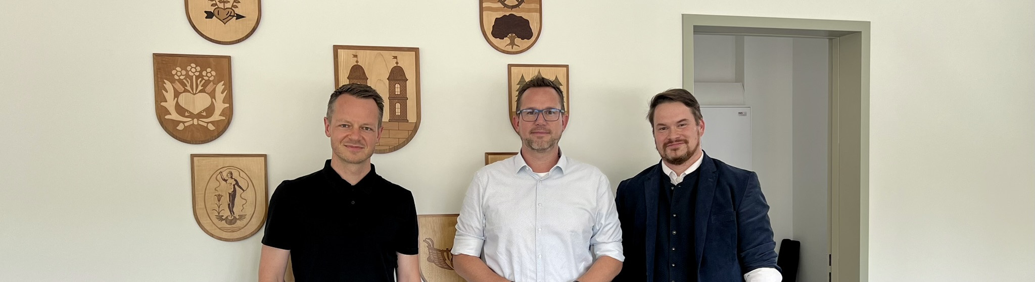 KI generiert: Das Bild zeigt drei Männer in einem Raum mit mehreren Holzschildern an der Wand, die verschiedene Symbole und Embleme darstellen. Die Personen stehen nebeneinander und posieren für das Foto.