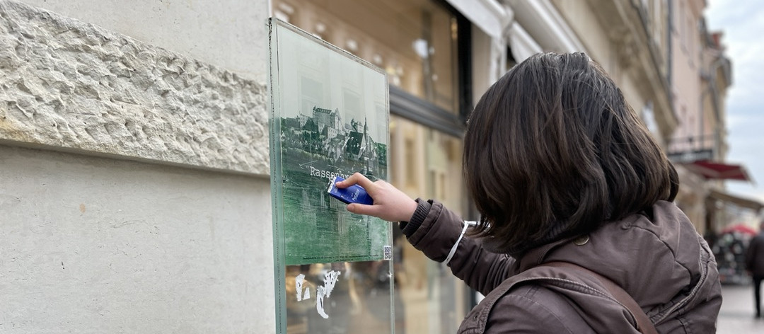KI generiert: Das Bild zeigt eine Person von hinten, die mit einem Handy gegen eine Glasscheibe klopft, auf der ein Bild einer Burg oder eines Schlosses zu sehen ist. Der Hauptinhalt des Bildes ist die Interaktion der Person mit dem Bild auf der Glasscheibe.