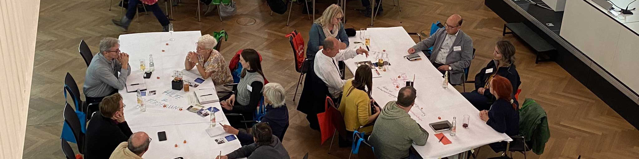 KI generiert: Das Bild zeigt eine Gruppe von Menschen, die an langen, mit Papier und Stiften bedeckten Tischen sitzen und sich in einer hellen, geräumigen Halle unterhalten. Der Hauptinhalt des Bildes ist eine Besprechung oder ein Workshop, bei dem die Teilnehmer gemeinsam arbeiten und diskutieren.