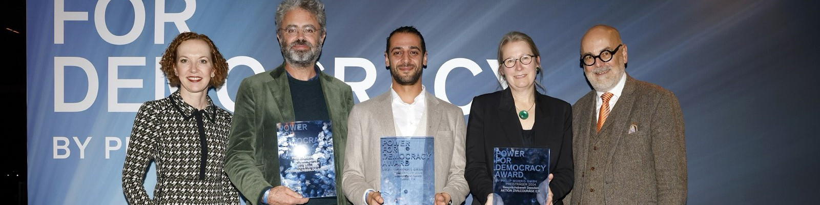 KI generiert: Das Bild zeigt fünf Personen, die auf einer Bühne stehen und jeweils eine Urkunde oder einen Preis in den Händen halten. Der Hintergrund deutet auf eine Veranstaltung hin, bei der ein "Democracy Award" verliehen wird.