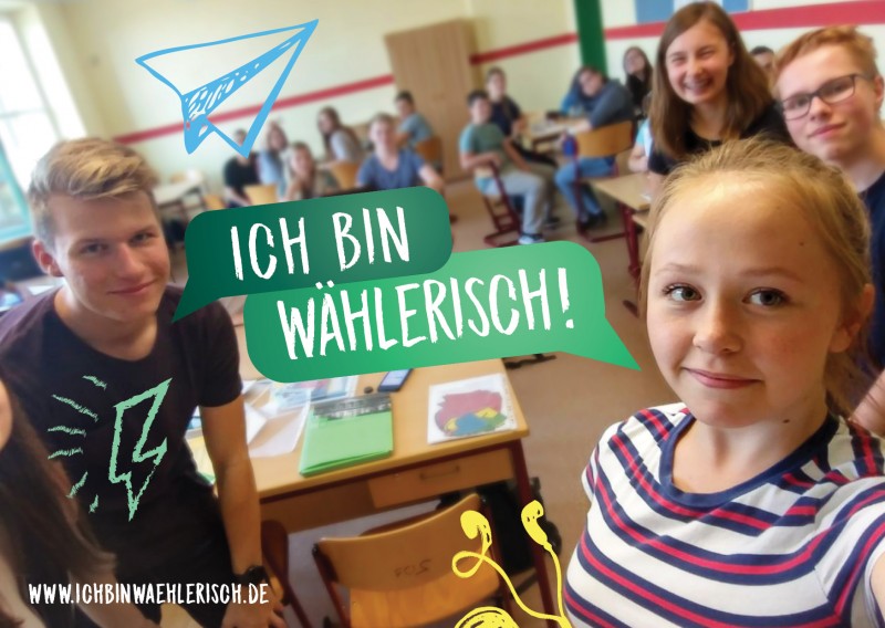 KI generiert: Das Bild zeigt eine Gruppe von Schülern in einem Klassenzimmer, wobei zwei Schüler im Vordergrund posieren und ein Text "ICH BIN WÄHLERISCH!" eingeblendet ist. Der Hauptinhalt des Bildes ist die Förderung der Wahlbeteiligung, insbesondere bei jungen Menschen.