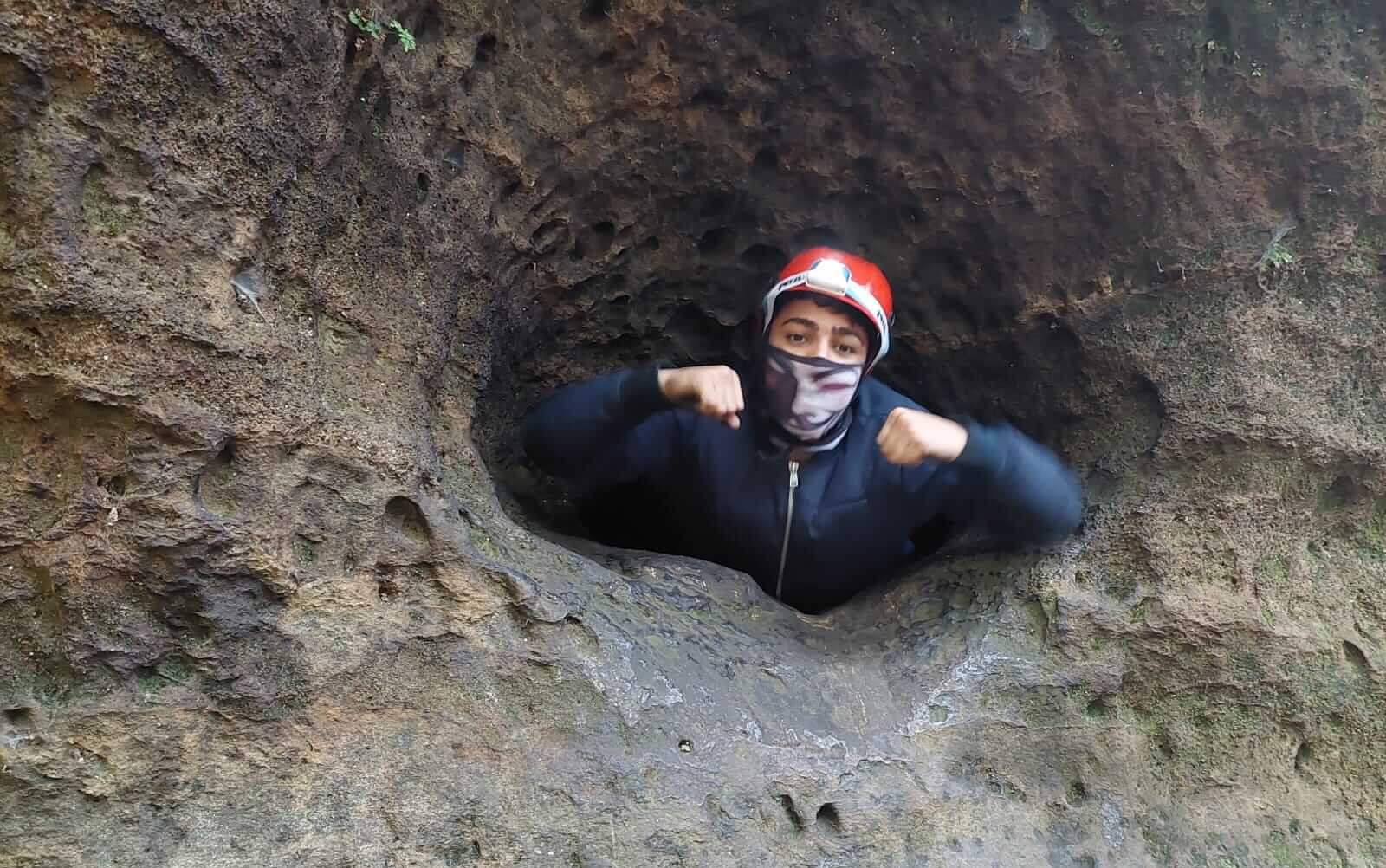 KI generiert: Das Bild zeigt eine Person, die durch ein enges Loch in einer Felswand klettert. Die Person trägt eine Jacke, einen roten Helm und eine Gesichtsmaske.