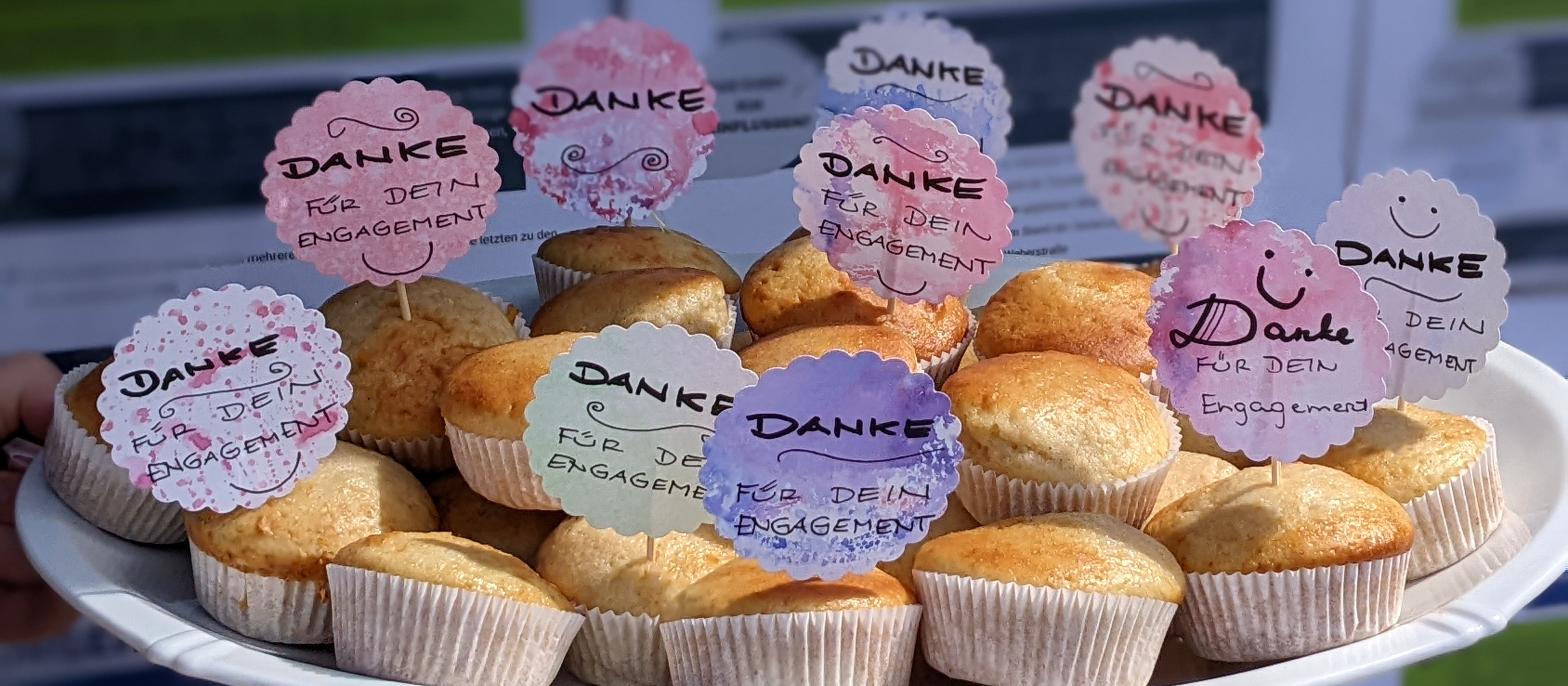 KI generiert: Das Bild zeigt eine Platte mit Muffins, die mit kleinen Schildern verziert sind, auf denen "Danke für dein Engagement" steht. Der Hauptinhalt des Bildes sind die Muffins und die Dankesbotschaften.