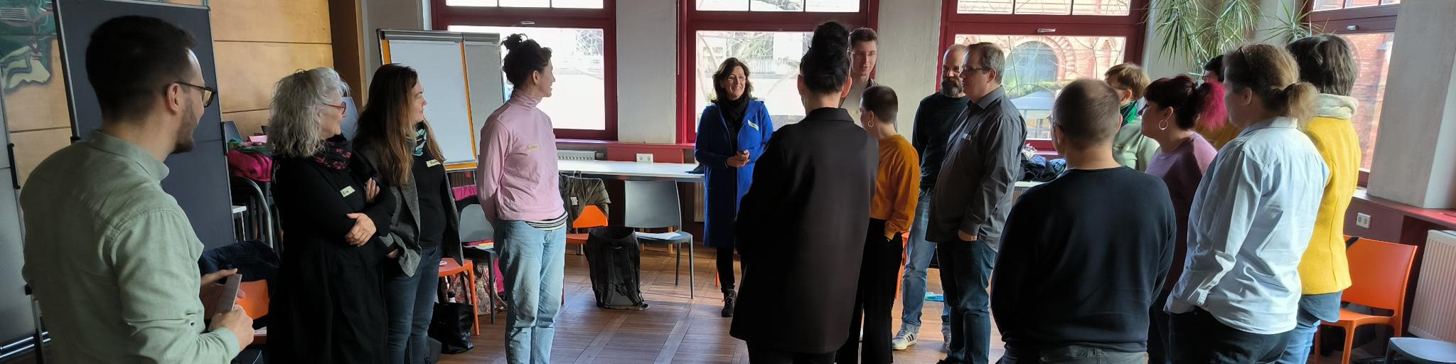 KI generiert: Das Bild zeigt eine Gruppe von Menschen, die in einem hellen Raum mit großen Fenstern stehen und sich unterhalten. Der Hauptinhalt des Bildes ist eine informelle Zusammenkunft oder ein Treffen, bei dem die Teilnehmer in kleinen Gruppen diskutieren.