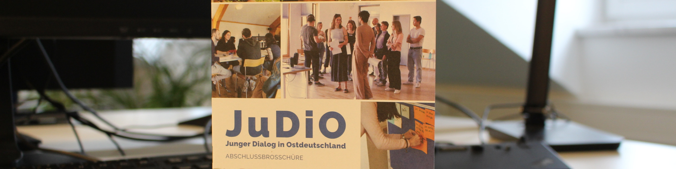 KI generiert: Das Bild zeigt eine Broschüre mit dem Titel "JuDiO - Junger Dialog in Ostdeutschland" auf einem Schreibtisch vor einem Computerbildschirm. Im Hintergrund sind Fotos von Menschen in Gesprächen und einer Person, die eine Präsentation hält, zu sehen.