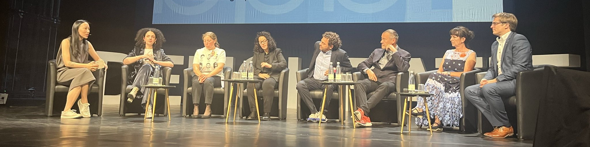 KI generiert: Das Bild zeigt eine Gruppe von acht Personen, die auf einer Bühne sitzen und an einer Diskussion oder einem Panel teilnehmen. Im Hintergrund ist eine große Leinwand mit einer blauen Grafik zu sehen.