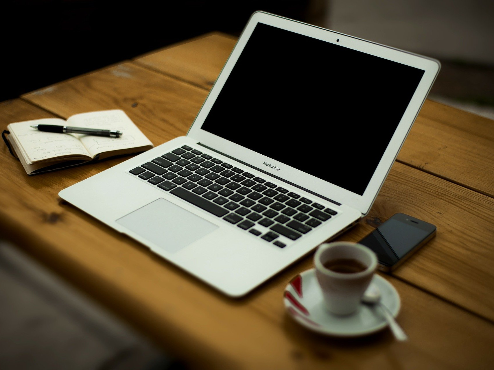 KI generiert: Das Bild zeigt einen Arbeitsplatz mit einem Laptop, einem Notizbuch, einem Stift, einem Smartphone und einer Tasse Kaffee auf einem Holztisch. Der Hauptinhalt des Bildes ist eine Arbeitsumgebung, die auf Produktivität und Konzentration ausgelegt ist.