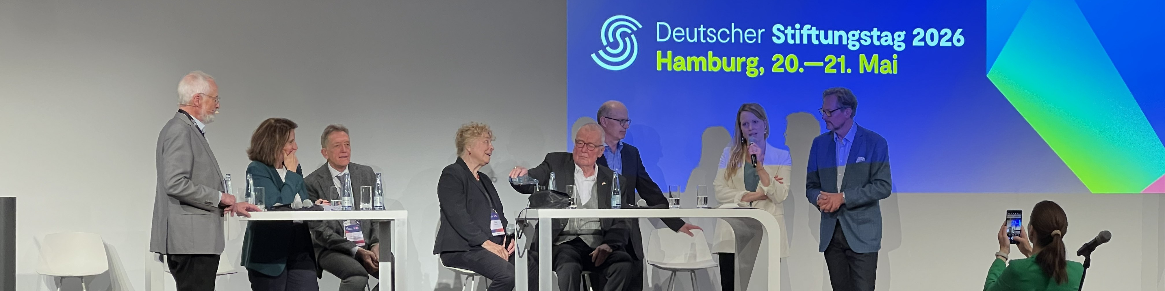 KI generiert: Das Bild zeigt eine Podiumsdiskussion mit mehreren Teilnehmern, die an Tischen sitzen und Mikrofone nutzen, während im Hintergrund eine Leinwand mit dem Titel "Deutscher Stiftungstag 2026, Hamburg, 20.–21. Mai" zu sehen ist. Der Hauptinhalt des Bildes ist die Darstellung einer Konferenz oder eines Fachtreffens, bei dem Experten oder Interessierte über Themen im Zusammenhang mit Stiftungen diskutieren.