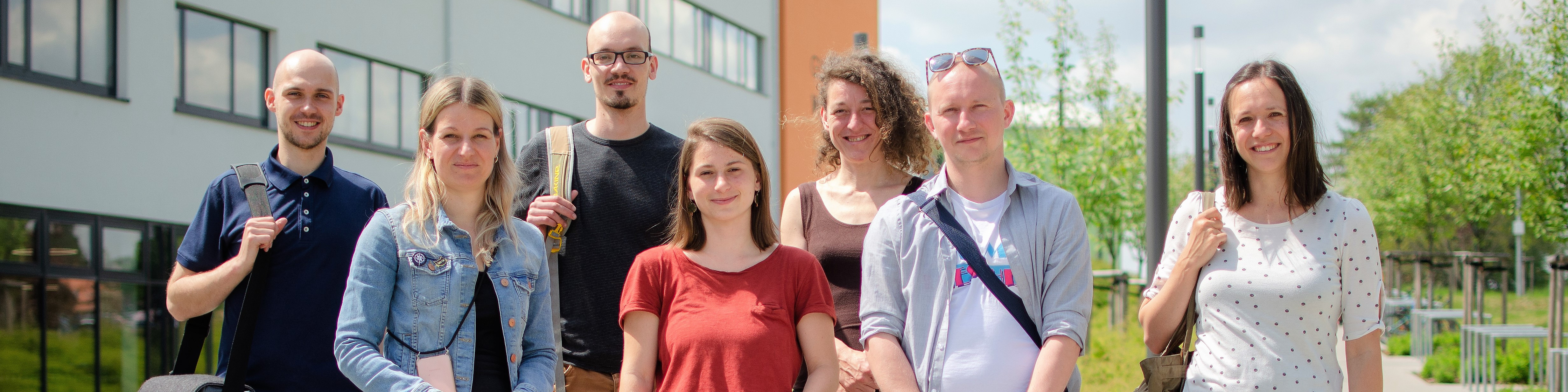 KI generiert: Das Bild zeigt eine Gruppe von sieben Personen, die vor einem Gebäude im Freien stehen und in die Kamera lächeln. Der Hauptinhalt des Bildes ist die fröhliche Gruppe, die sich möglicherweise zu einem Anlass oder einer Veranstaltung versammelt hat.