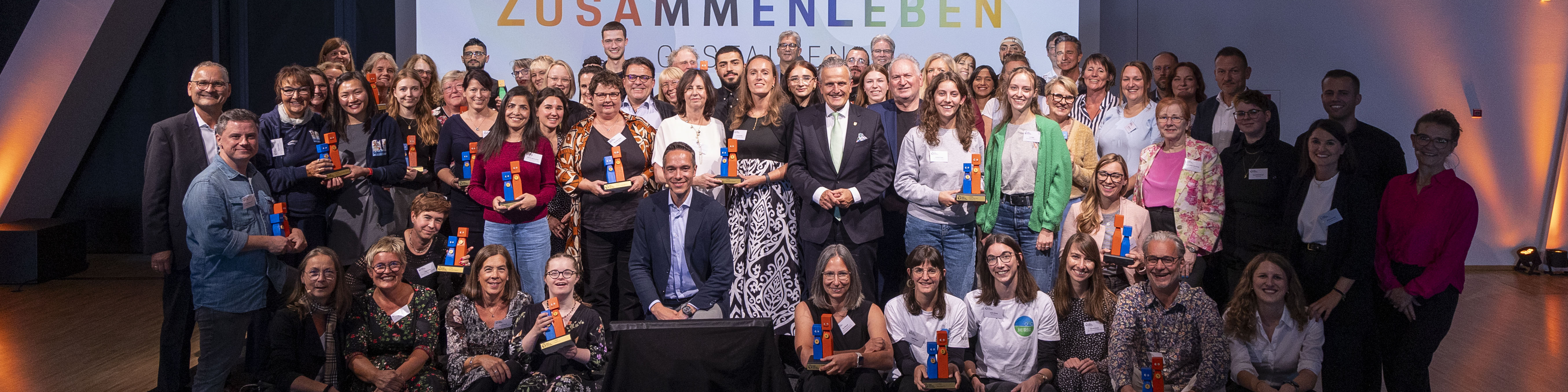 KI generiert: Das Bild zeigt eine große Gruppe von Menschen, die auf einer Bühne stehen und gemeinsam posieren. Viele von ihnen halten eine orangefarbene Urkunde oder einen Gegenstand in der Hand, und im Hintergrund ist ein Banner mit der Aufschrift "ZUSAMMENLEBEN" zu sehen.