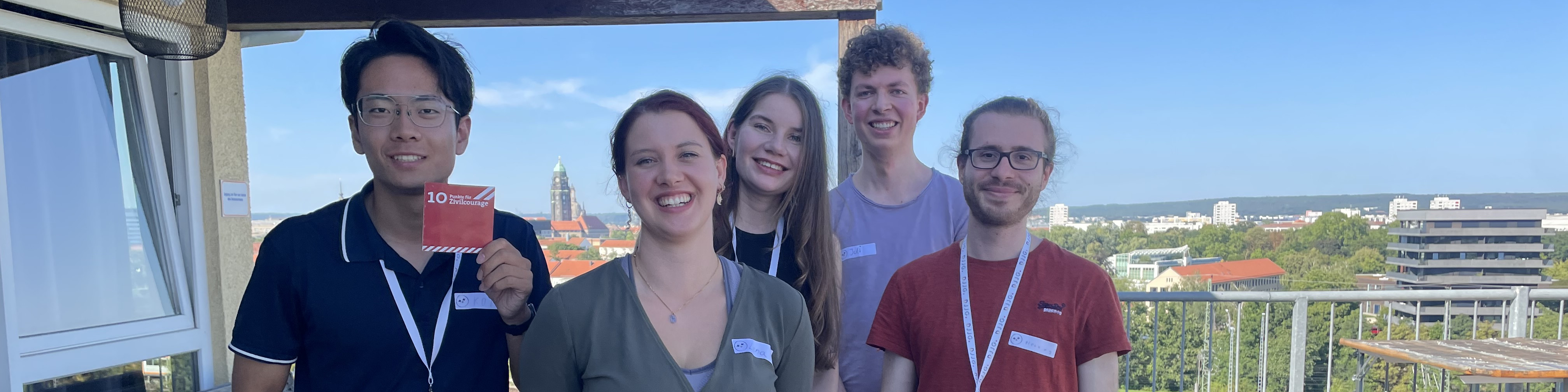 KI generiert: Das Bild zeigt eine Gruppe von fünf Personen, die auf einem Balkon stehen und in die Kamera lächeln. Im Hintergrund ist eine Stadtlandschaft mit Gebäuden und Bäumen zu sehen, und eine Person hält ein rotes Schild mit der Aufschrift "10 Jahre".
