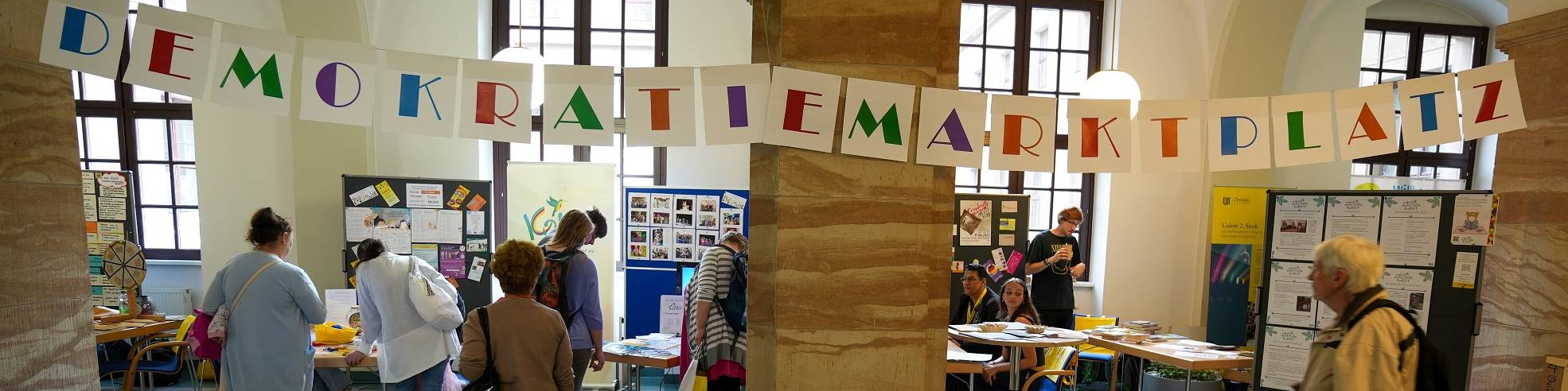 KI generiert: Das Bild zeigt eine Ausstellungs- oder Informationsveranstaltung in einem hellen, historischen Raum mit mehreren Ständen und Postern. Der Hauptinhalt ist der "DemokratieMarktPlatz", wie auf dem Banner über den Ständen zu lesen ist.