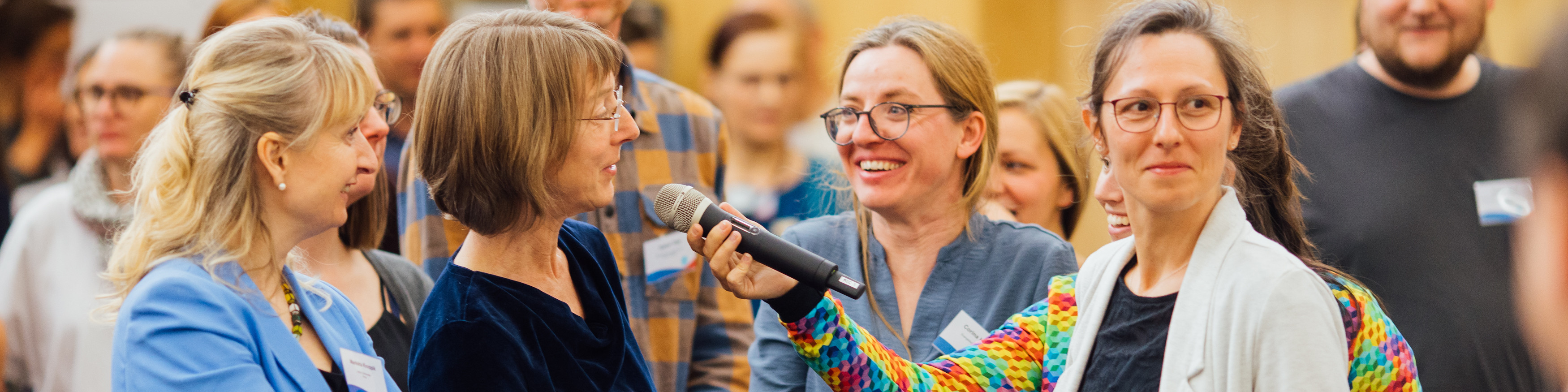 KI generiert: Das Bild zeigt eine Gruppe von Personen, die sich in einer Versammlung oder einem Seminar befinden, wobei eine Frau ein Mikrofon hält und möglicherweise spricht oder eine Frage stellt. Der Hauptinhalt des Bildes ist die Interaktion und Kommunikation zwischen den Teilnehmern in einer öffentlichen oder halboffentlichen Veranstaltung.