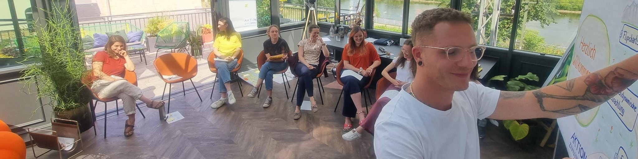 KI generiert: Das Bild zeigt eine Gruppe von Personen, die in einem hellen, modernen Raum mit großen Fenstern und Pflanzen sitzen und einer Präsentation folgen. Der Hauptinhalt ist eine Schulung oder ein Workshop, bei dem ein Mann vorne steht und etwas an einem Flipchart erklärt.