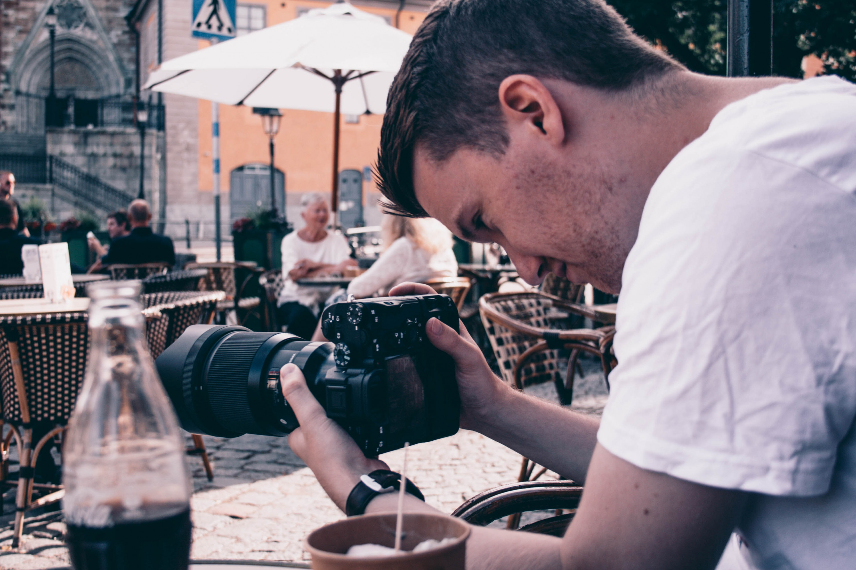 KI generiert: Das Bild zeigt einen Mann, der sich über einen Tisch beugt und eine Kamera mit einem großen Teleobjektiv bedient. Im Vordergrund steht ein Glas mit einem dunklen Getränk, möglicherweise Cola, und im Hintergrund sind Menschen an Tischen in einem Café oder einer öffentlichen Platzsituation zu sehen. Der Hauptinhalt des Bildes ist die Fotografie und die entspannte Atmosphäre eines öffentlichen Ortes.