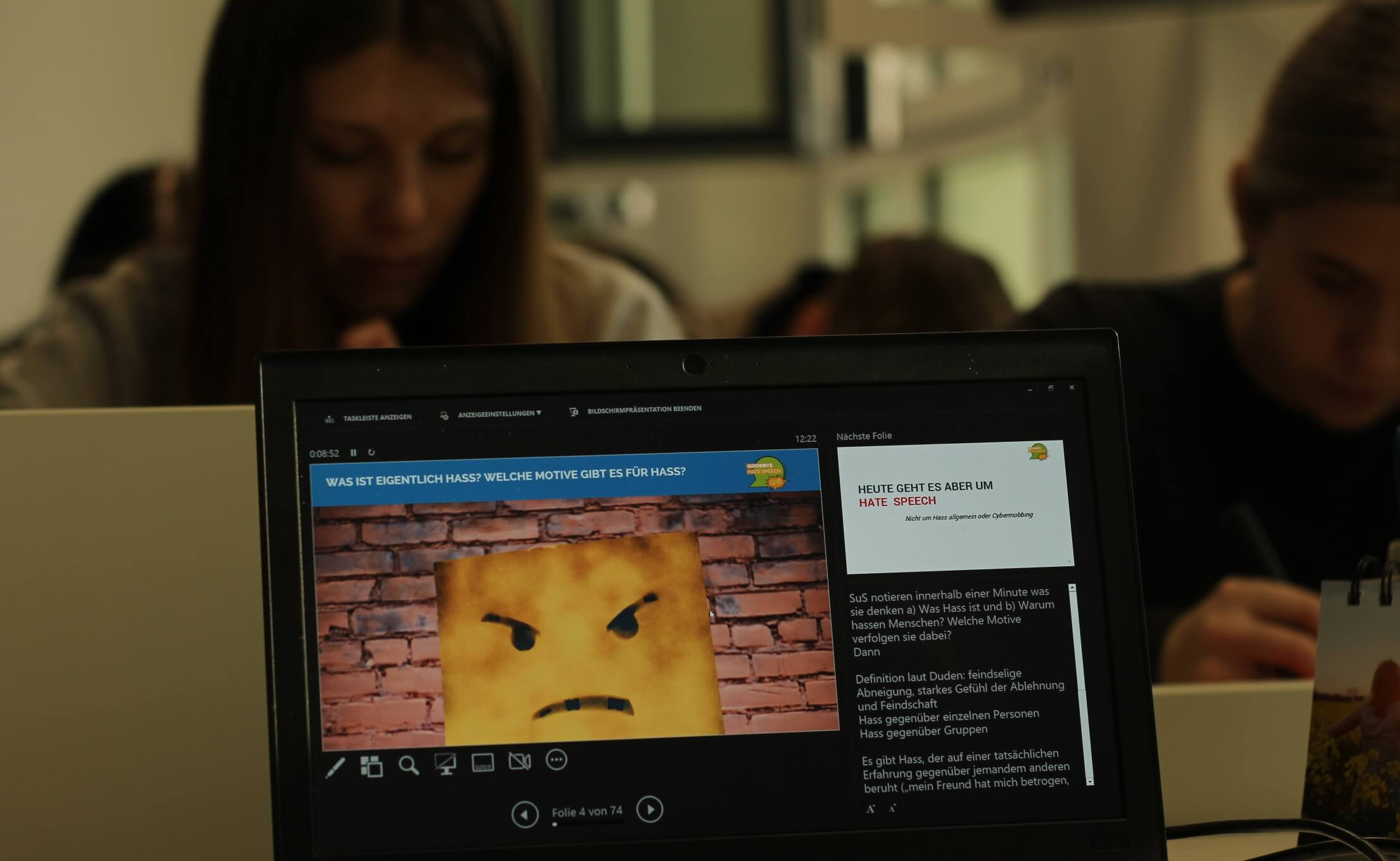 KI generiert: Das Bild zeigt eine Person, die auf einen Laptop schaut, auf dessen Bildschirm eine Präsentation oder ein Artikel mit dem Titel "Was ist eigentlich Hass? Welche Motive gibt es für Hass?" zu sehen ist. Im Vordergrund ist ein Bild mit einem wütenden Emoji vor einer Backsteinmauer zu erkennen.