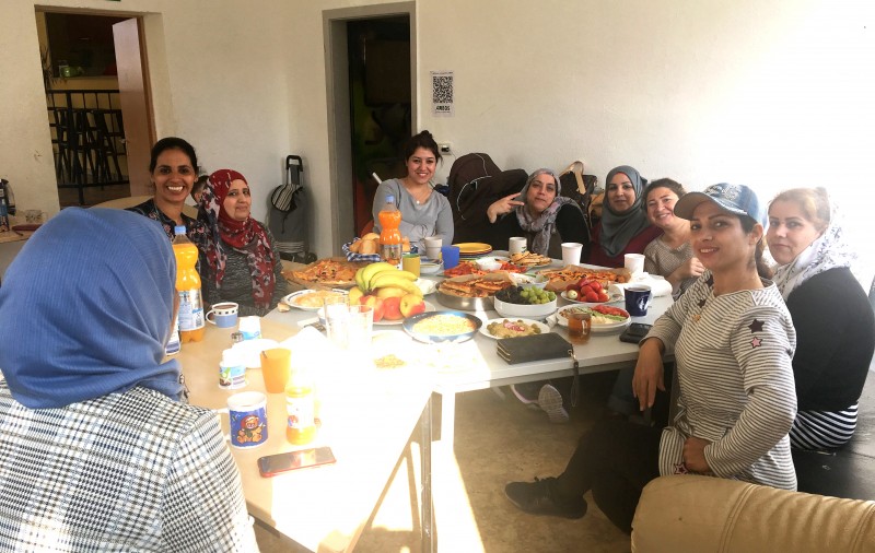 KI generiert: Das Bild zeigt eine Gruppe von Frauen, die gemeinsam an einem langen Tisch sitzen und ein Essen teilen. Der Tisch ist mit verschiedenen Speisen, Getränken und Obst beladen, was auf eine gesellige Zusammenkunft hinweist.