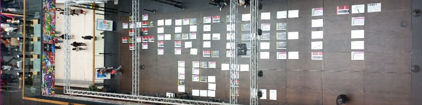 KI generiert: Das Bild zeigt eine Wand mit vielen kleinen, an Pinnwänden befestigten Zetteln oder Karten, die in einem strukturierten Raster angeordnet sind. Im Vordergrund sind Regale mit verschiedenen Gegenständen zu sehen, darunter bunte Kabel und technische Ausrüstung.