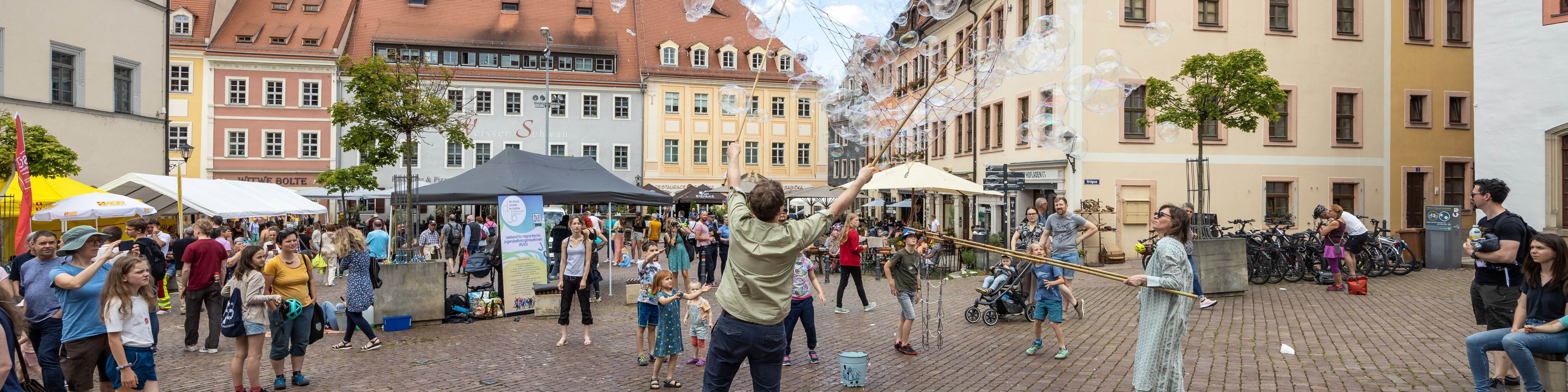 KI generiert: Das Bild zeigt eine belebte Platzszene mit Menschen, die eine Seifenblasen-Show beobachten, während im Hintergrund bunte Gebäude und Marktstände zu sehen sind. Der Hauptinhalt ist die unterhaltsame Atmosphäre eines Straßenfestes oder einer öffentlichen Veranstaltung.