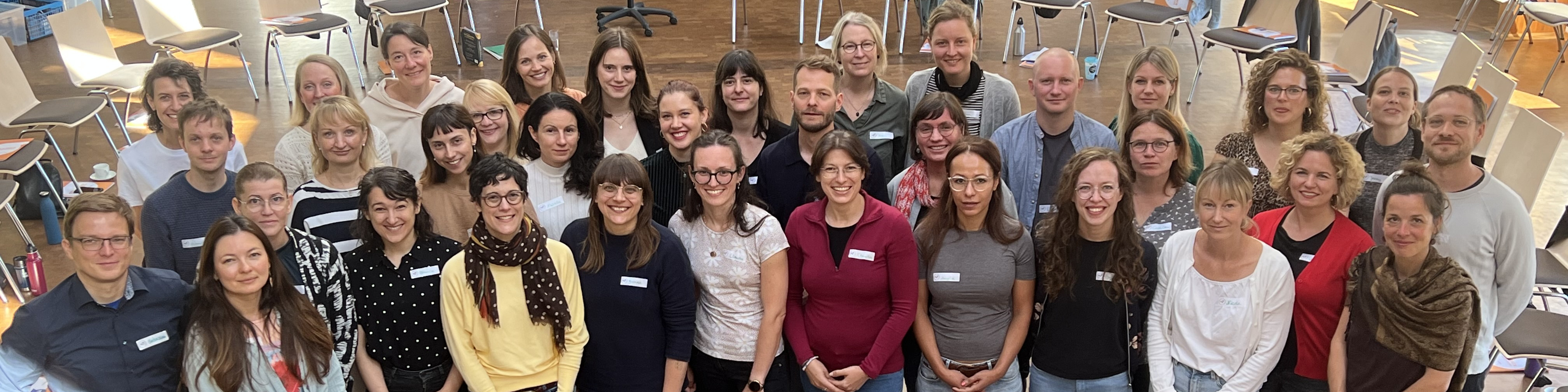 KI generiert: Das Bild zeigt eine große Gruppe von Menschen, die in einem hellen Raum mit Stühlen und Tischen posieren. Der Hauptinhalt des Bildes ist eine Gruppenfotografie, möglicherweise von einer Konferenz oder einem Workshop.