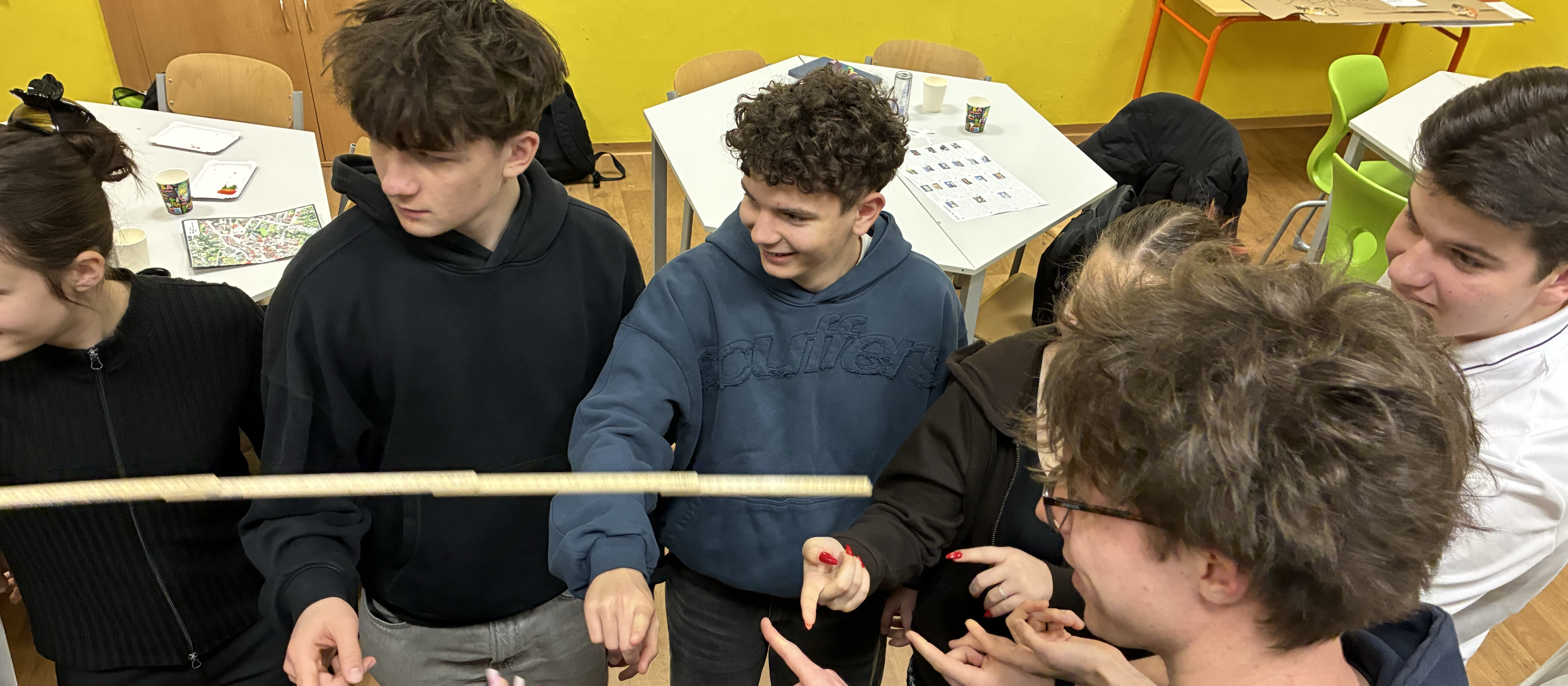 KI generiert: Das Bild zeigt eine Gruppe junger Menschen, die sich um einen Tisch versammelt haben und gemeinsam eine Aktivität oder ein Spiel durchführen. Der Hauptinhalt des Bildes ist die Interaktion und Zusammenarbeit der Gruppe in einer scheinbar lockeren und geselligen Atmosphäre.