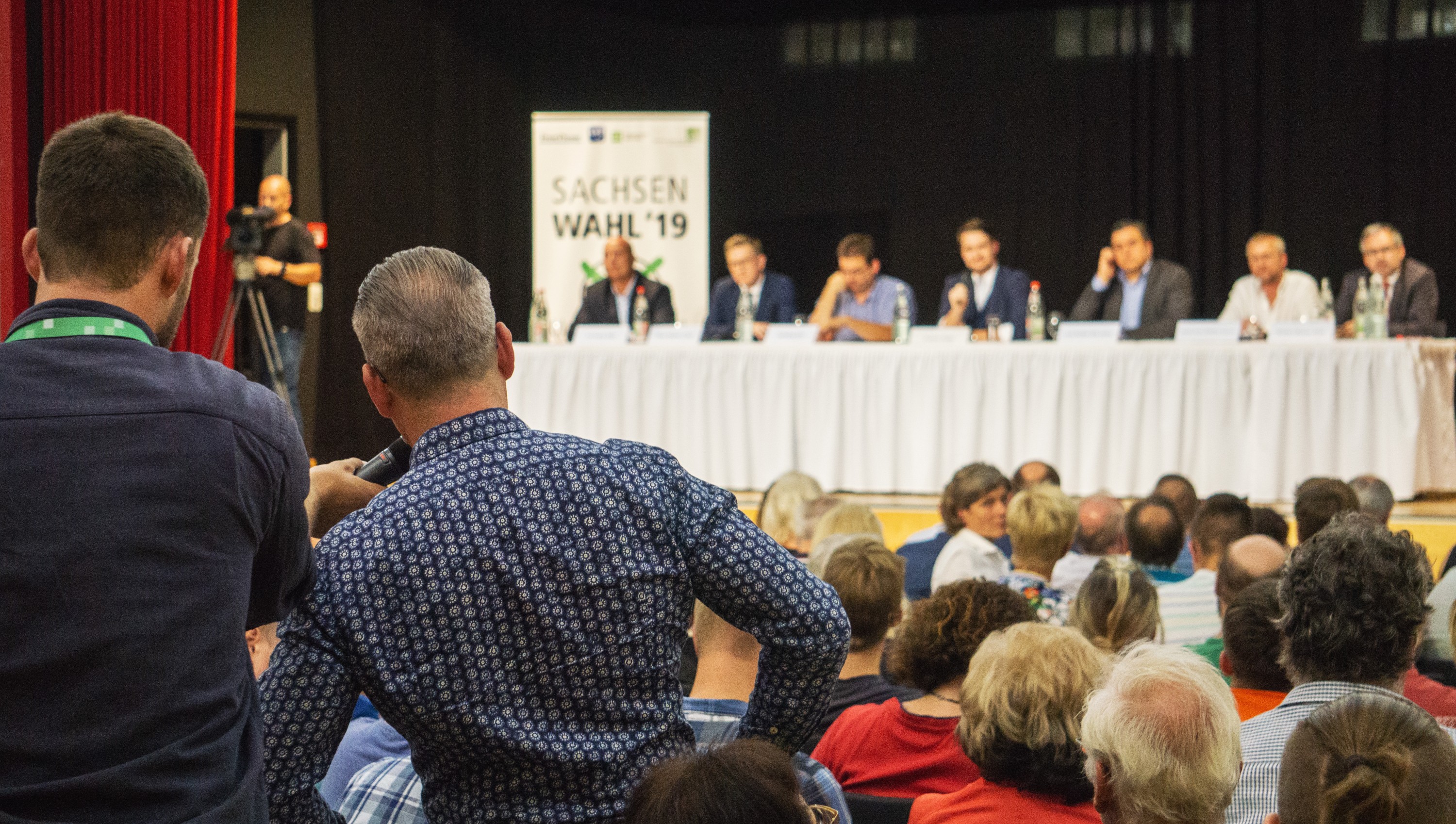 KI generiert: Das Bild zeigt eine öffentliche Veranstaltung oder Podiumsdiskussion mit mehreren Teilnehmern auf der Bühne, die vor einem Publikum sprechen. Im Hintergrund ist ein Banner mit der Aufschrift "SACHSEN WAHL '19" zu sehen, was darauf hindeutet, dass es sich um eine Wahlveranstaltung in Sachsen handelt.