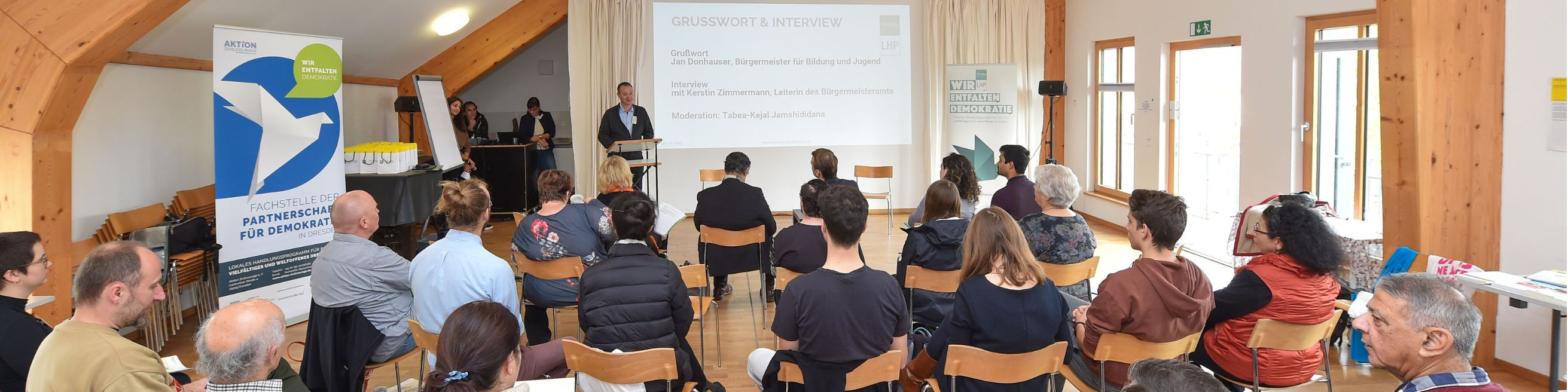 KI generiert: Das Bild zeigt eine Versammlung oder Konferenz in einem hellen, holzvertäfelten Raum, bei der eine Person vorne steht und zu den Anwesenden spricht, während eine Präsentation auf einem Bildschirm im Hintergrund läuft. Der Hauptinhalt des Bildes ist eine formelle Präsentation oder Diskussion in einer Gruppenumgebung.
