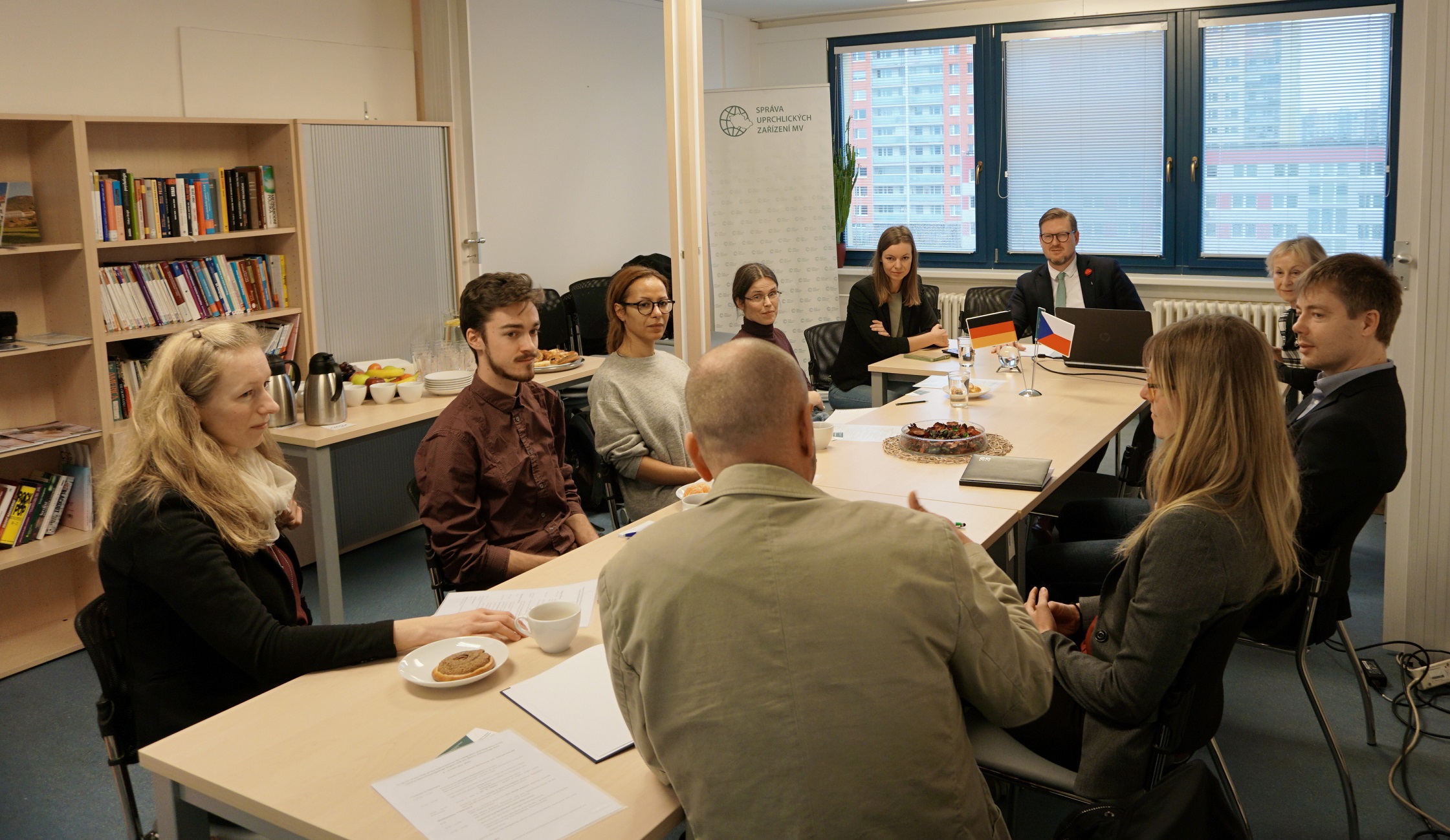 KI generiert: Das Bild zeigt eine Gruppe von Personen, die in einem Besprechungsraum an einem langen Tisch sitzen und sich unterhalten. Im Vordergrund sind einige Teilnehmer im Gespräch, während im Hintergrund weitere Personen am Tisch sitzen, darunter eine Person mit kleinen Flaggen vor sich.