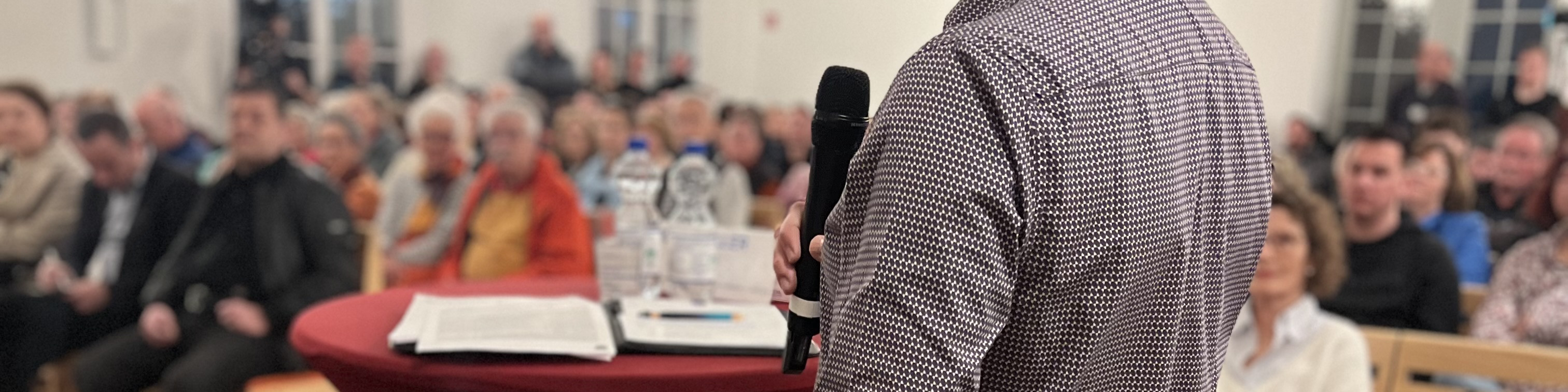 KI generiert: Das Bild zeigt eine Person, die vor einem Publikum spricht und dabei ein Mikrofon hält. Im Vordergrund sind ein Tisch mit einer Flasche Wasser, einem Glas und einigen Papieren zu sehen.
