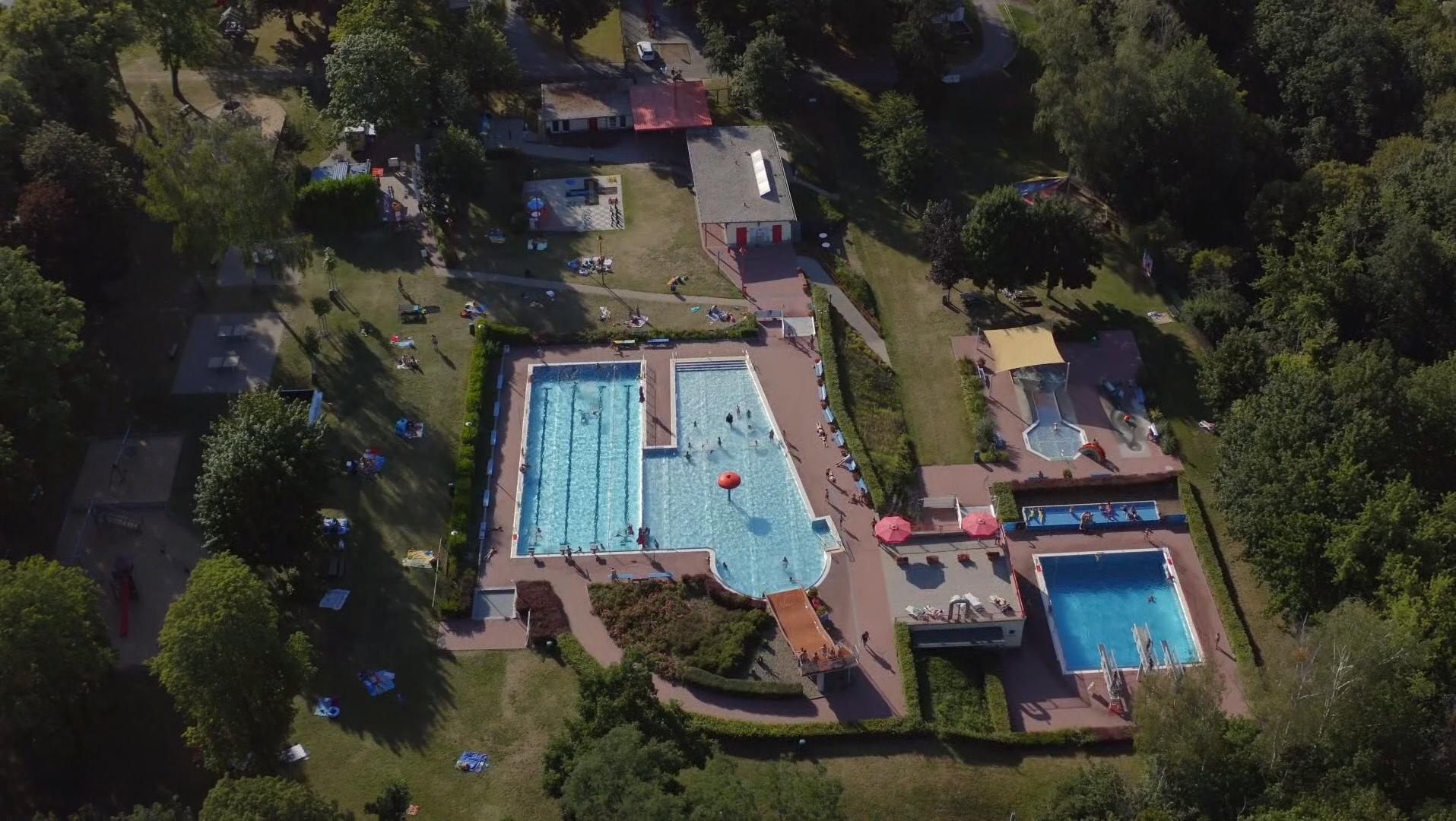 KI generiert: Das Bild zeigt eine Luftaufnahme eines Freizeitbades mit mehreren Schwimmbecken, umgeben von Grünflächen und Gebäuden. Der Hauptinhalt des Bildes ist das Schwimmbad mit seinen verschiedenen Becken und den umliegenden Freizeiteinrichtungen.