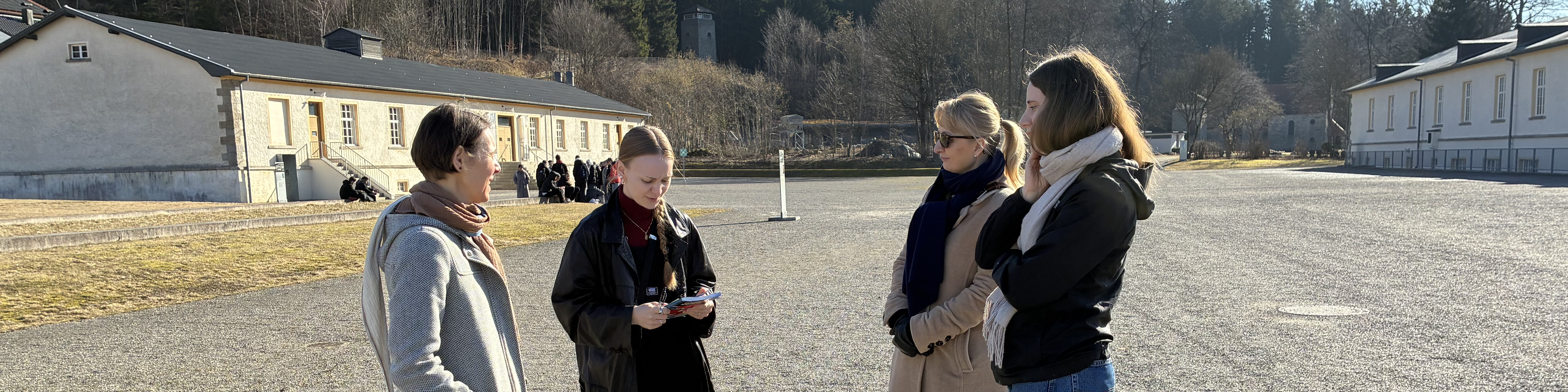 KI generiert: Das Bild zeigt eine Gruppe von vier Personen, die sich auf einem großen, offenen Platz unterhalten, während sie auf ein Smartphone schauen. Im Hintergrund sind Gebäude und eine Gruppe von Menschen zu sehen, die möglicherweise eine Führung oder Besichtigung machen.