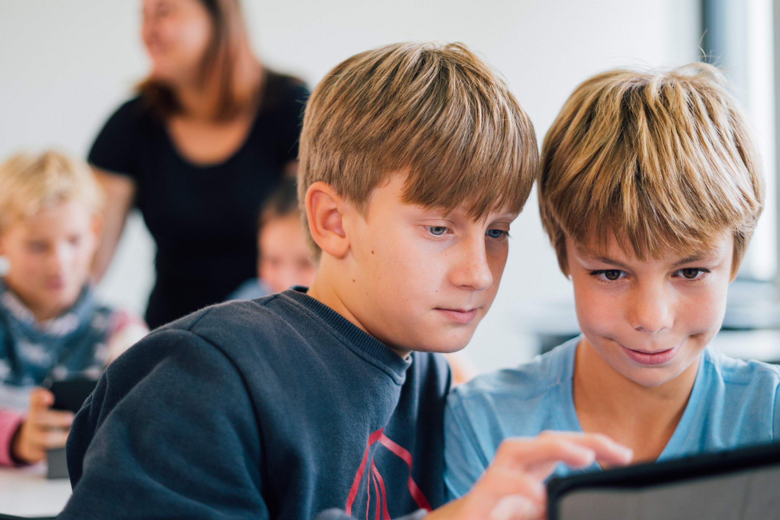 KI generiert: Das Bild zeigt zwei Jungen, die gemeinsam auf ein Tablet schauen, während sie in einem Klassenzimmer sitzen. Im Hintergrund sind weitere Kinder und eine erwachsene Person zu erkennen.