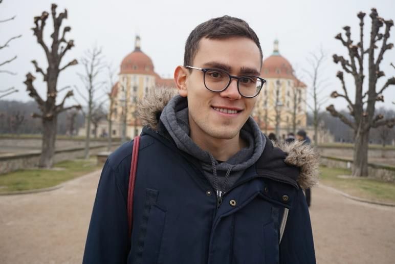 KI generiert: Das Bild zeigt einen lächelnden Mann mit Brille und Winterjacke, der vor einem historischen Gebäude mit Kuppeln und barocken Architekturmerkmalen steht. Im Hintergrund sind kahl geschnittene Bäume und ein Weg zu sehen.