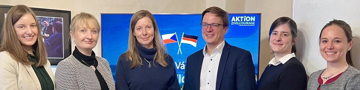 KI generiert: Das Bild zeigt eine Gruppe von sechs Personen, die vor einem Banner mit der Aufschrift "AKTION Zivilcourage" und den Flaggen von Tschechien und Deutschland stehen. Der Hauptinhalt des Bildes ist die Darstellung einer Zusammenarbeit oder eines Treffens im Rahmen einer Initiative für Zivilcourage.