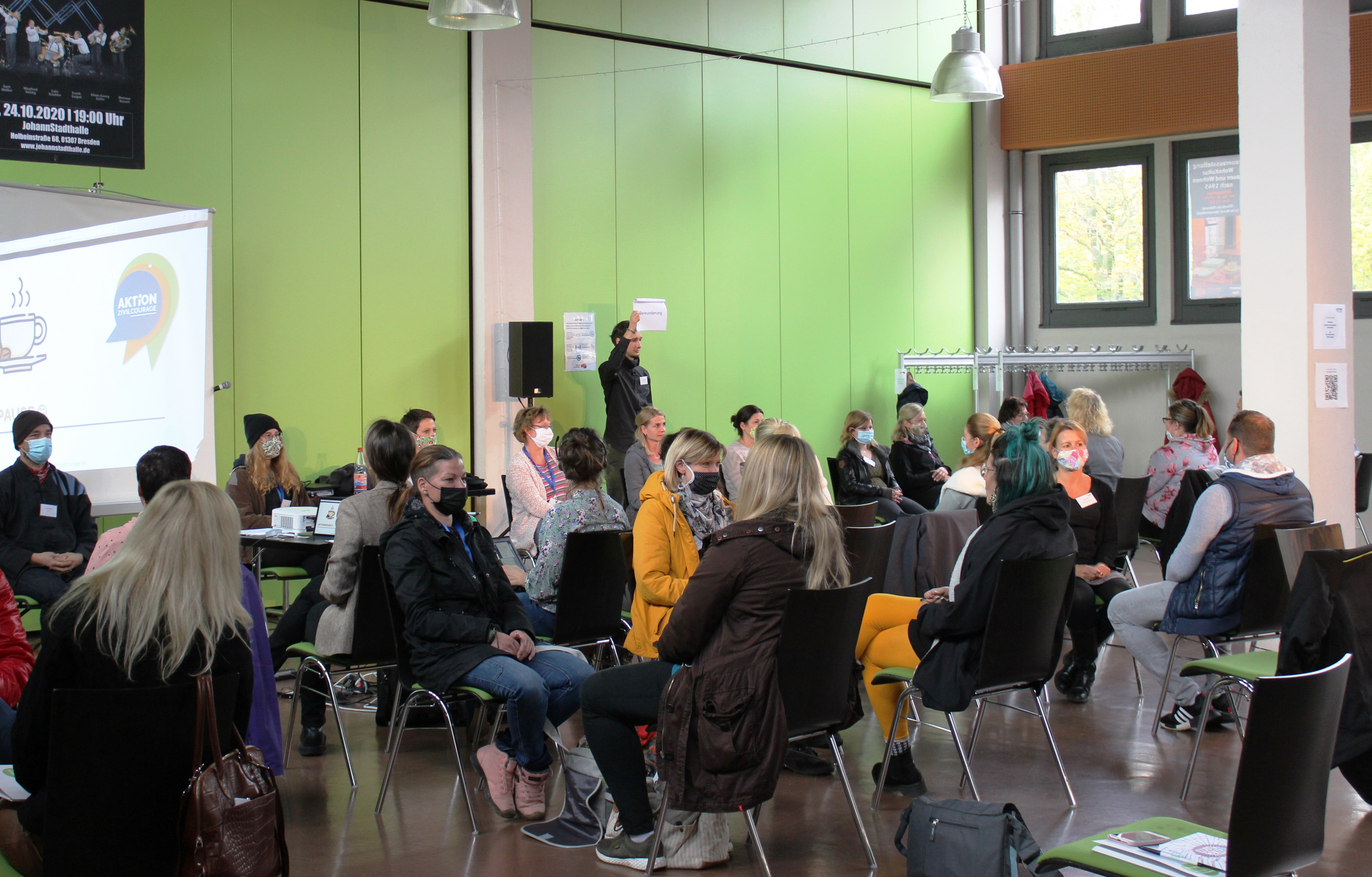 KI generiert: Das Bild zeigt eine Gruppe von Personen, die in einem hellen Raum mit grünen Wänden an einer Veranstaltung teilnehmen. Der Hauptinhalt ist eine Präsentation oder ein Workshop, bei dem eine Person vorne steht und zu den Anwesenden spricht.