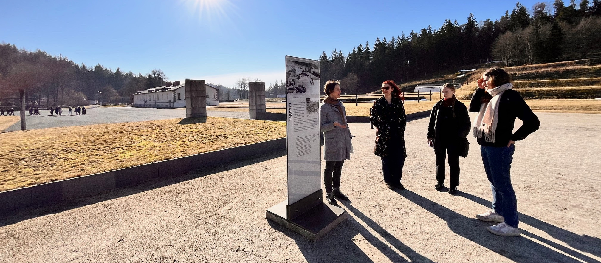 KI generiert: Das Bild zeigt eine Gruppe von vier Personen, die eine Informationsplatte in einem Freiluftbereich betrachten. Im Hintergrund sind historische Gebäude und eine weitläufige, offene Landschaft zu sehen, die auf einen Ort von historischem Interesse hindeuten könnte.