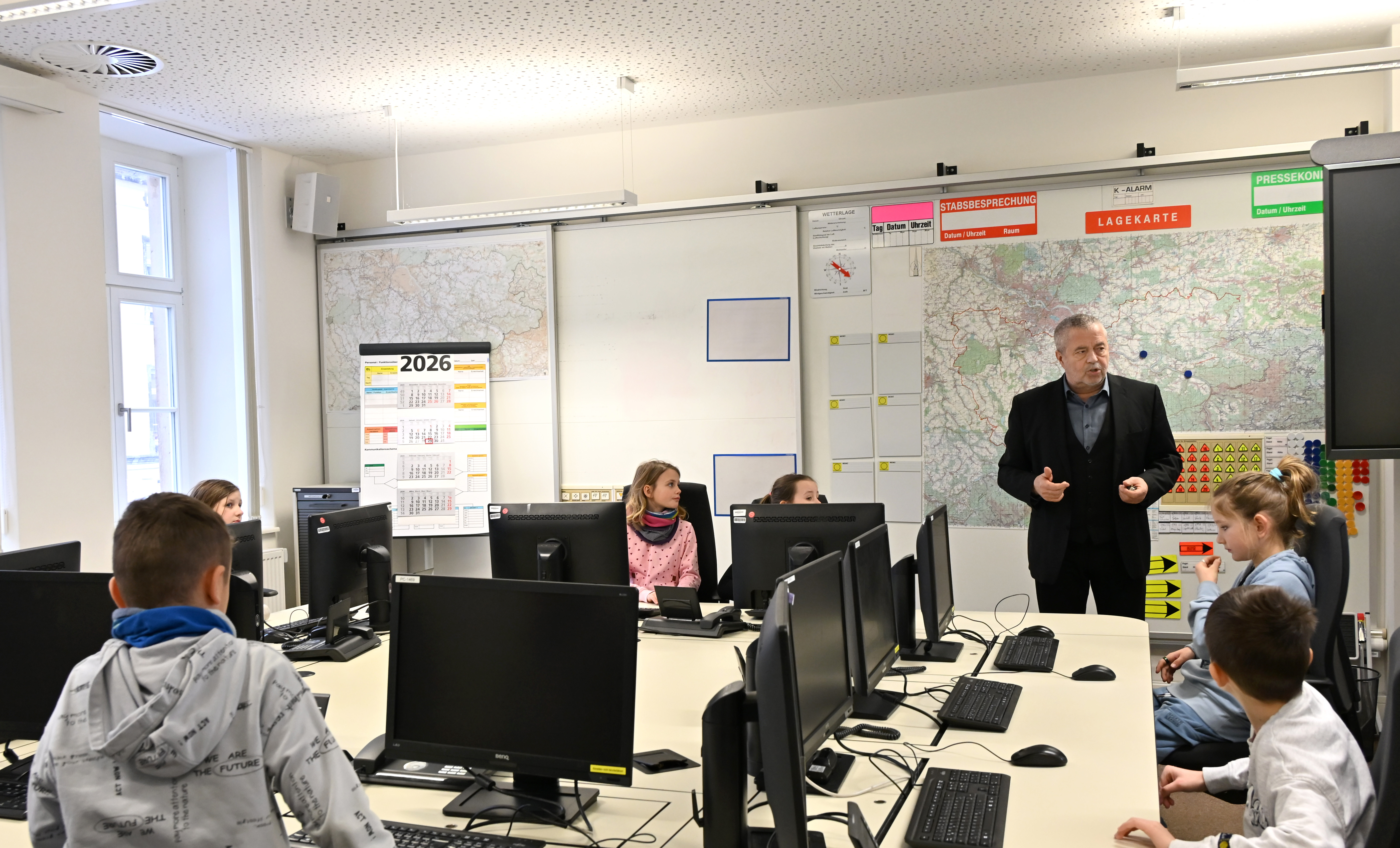KI generiert: Das Bild zeigt einen Klassenraum mit Schülern, die an Computern arbeiten, während ein Lehrer vorne steht und mit ihnen spricht. Der Raum ist mit verschiedenen Karten und einem Kalender für das Jahr 2026 ausgestattet.