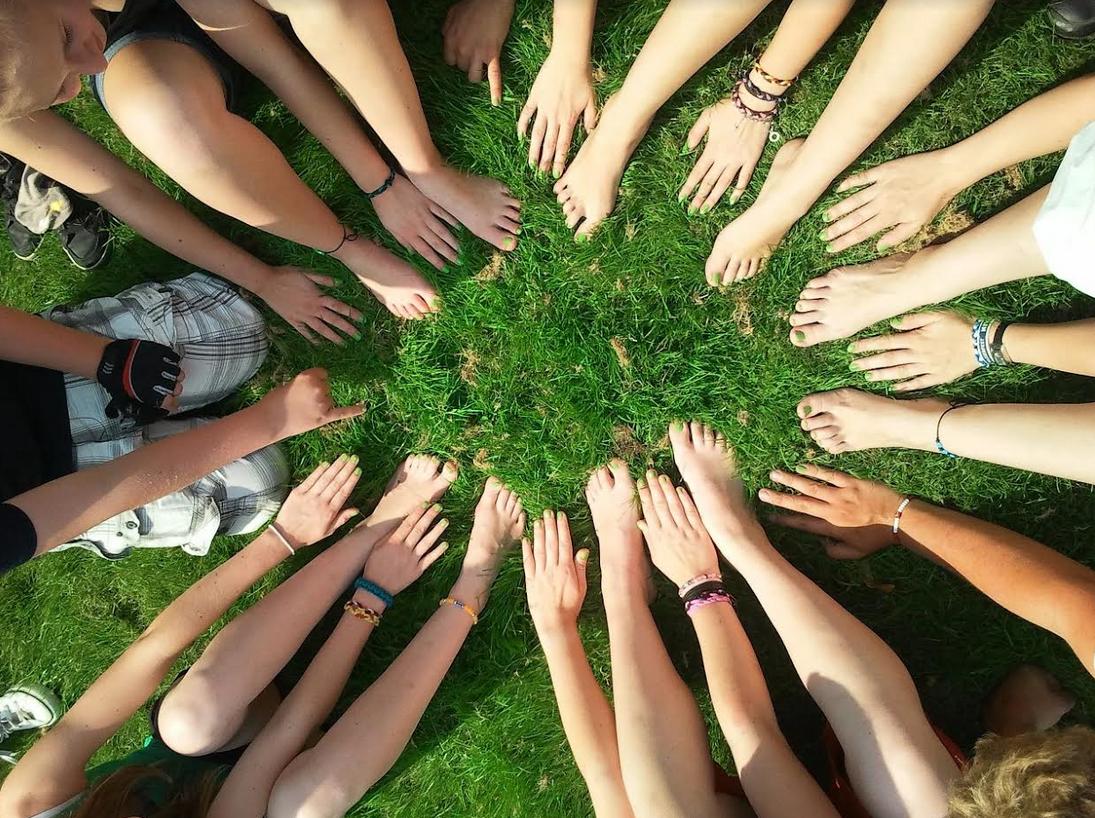 KI generiert: Das Bild zeigt eine Gruppe von Menschen, die im Kreis auf dem Boden sitzen und ihre Hände in die Mitte legen, wobei ihre Füße ebenfalls im Kreis angeordnet sind. Der Hauptinhalt des Bildes ist die Darstellung von Teamarbeit und Zusammenhalt.