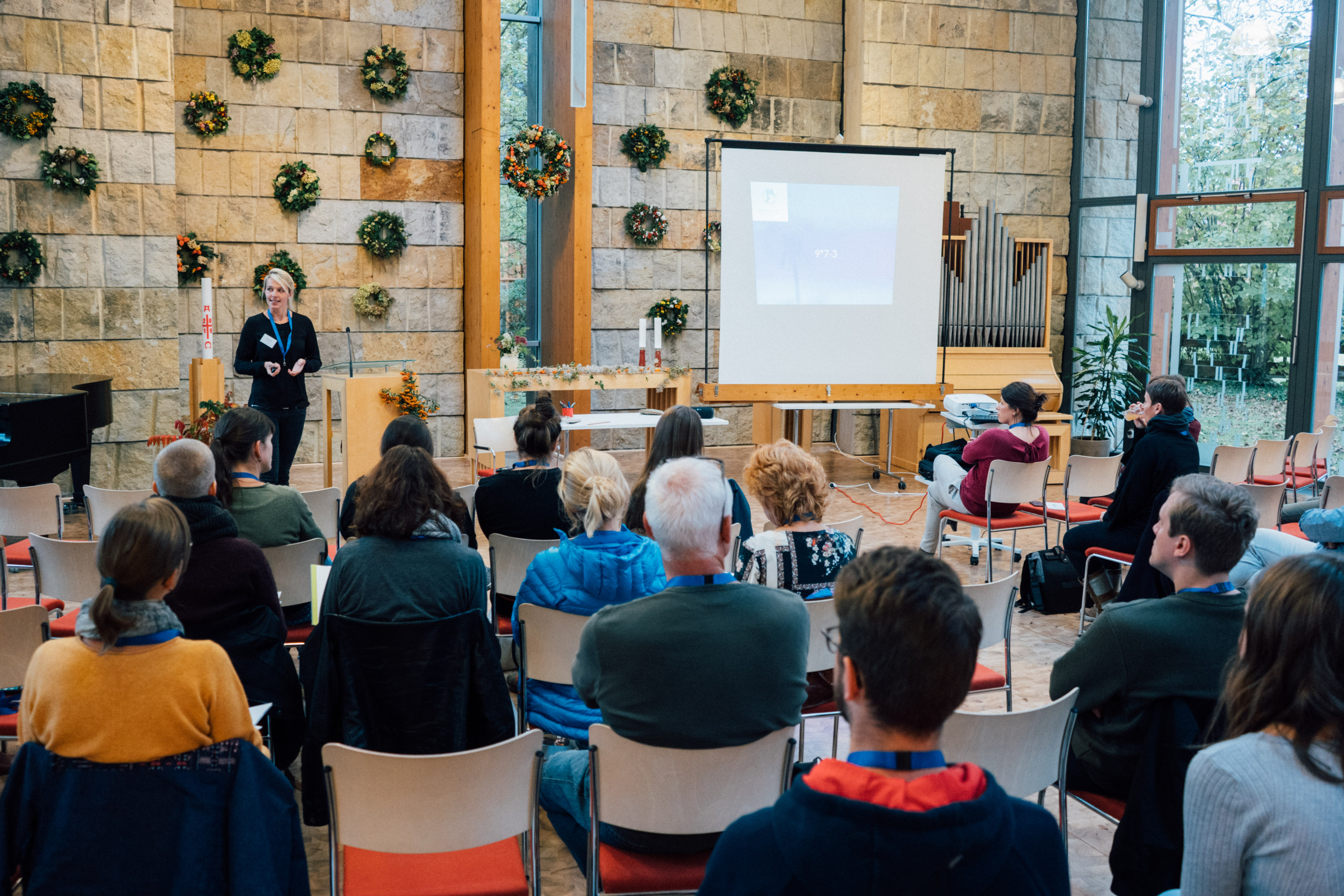 KI generiert: Das Bild zeigt eine Gruppe von Menschen, die in einem Raum mit Steinwänden und großen Fenstern sitzen und einer Person zuhören, die vorne steht und eine Präsentation hält. Der Hauptinhalt des Bildes ist eine Vortrags- oder Seminarveranstaltung.