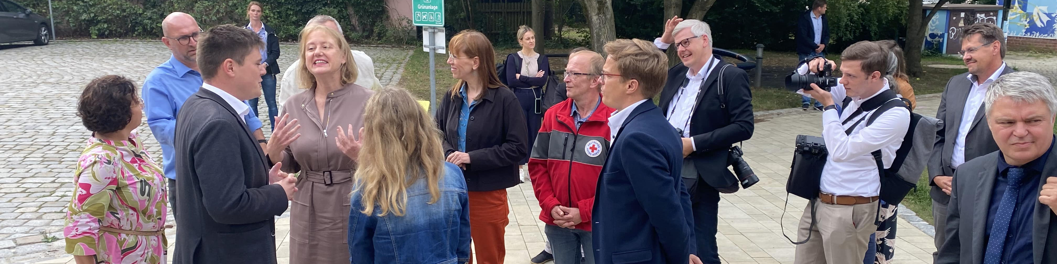 KI generiert: Das Bild zeigt eine Gruppe von Personen, die sich im Freien auf einem gepflasterten Platz unterhalten. Im Vordergrund stehen zwei Personen im Gespräch, während im Hintergrund weitere Menschen in kleinen Gruppen zusammenstehen und sich unterhalten.