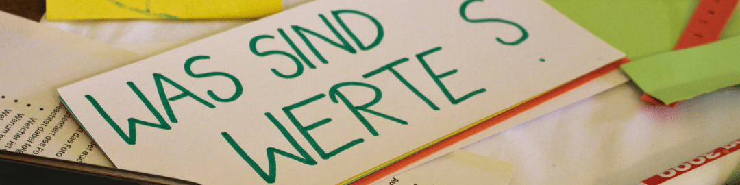 KI generiert: Das Bild zeigt ein Blatt Papier mit der handschriftlichen Aufschrift "WAS SIND WERTE S". Im Hintergrund sind weitere Papiere und ein grüner Ordner zu sehen.