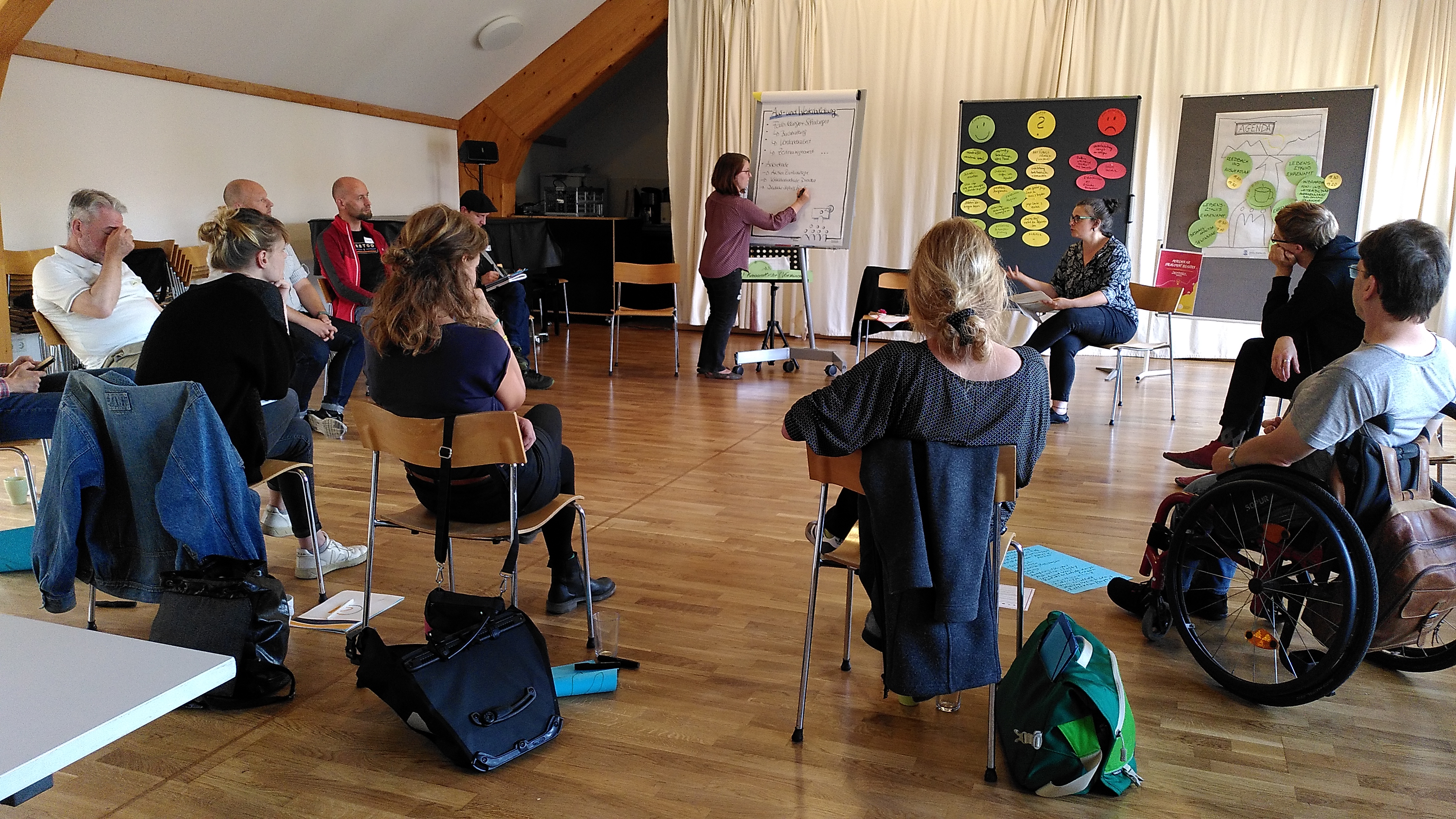 KI generiert: Das Bild zeigt eine Gruppe von Menschen, die in einem hellen Raum mit Holzfußboden an einem Seminar oder Workshop teilnehmen. Eine Person steht vorne und präsentiert Inhalte an einem Flipchart, während die anderen Teilnehmer auf Stühlen sitzen und zuhören.