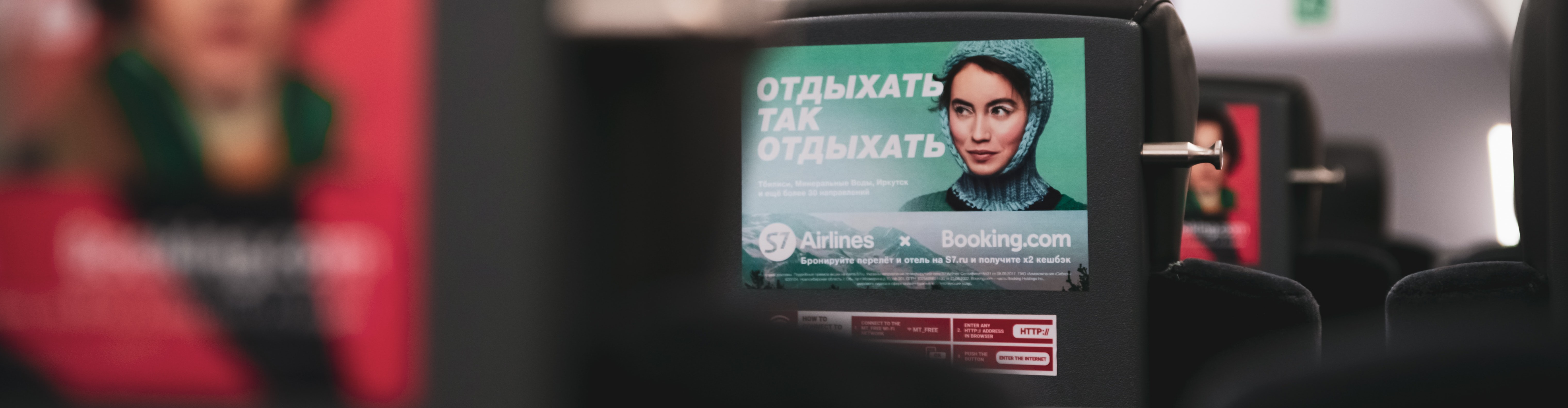 KI generiert: Das Bild zeigt die Rückenlehne eines Flugzeugsitzes mit einer Werbeanzeige, die eine lächelnde Person und russischen Text zeigt. Die Anzeige wirbt für eine Fluggesellschaft und eine Buchungswebsite.